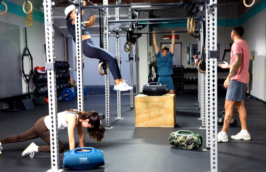 Lidé v posilovně, poslouchající hudbu z přenosného reproduktoru JBL Boombox 4